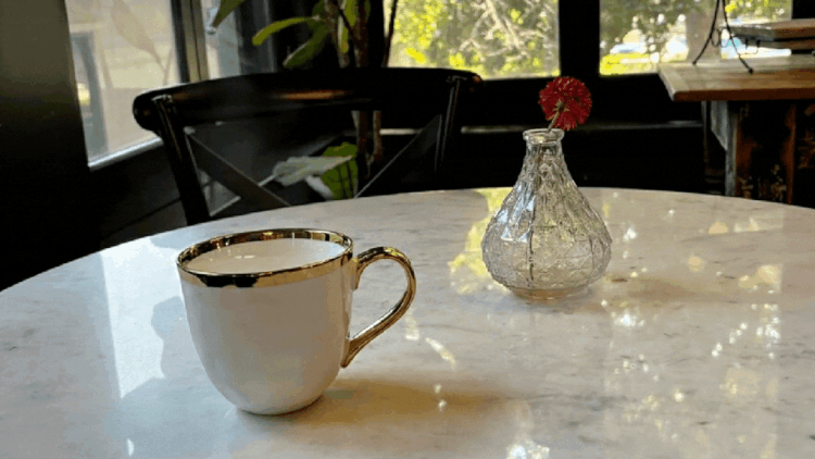 A white ceramic coffee cup with gold trim on a white marble table, next to a small glass vase with a red flower, near a window with green trees visible outside.
